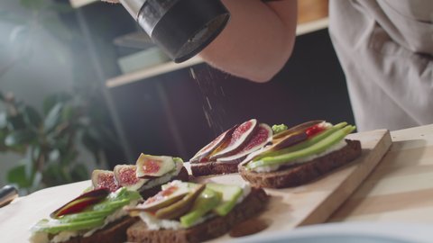 Dutch angle close up shot of hands of unrecognizable man adding black pepper from electric grinder to gourmet toasts with cream cheese, fig and avocado slices