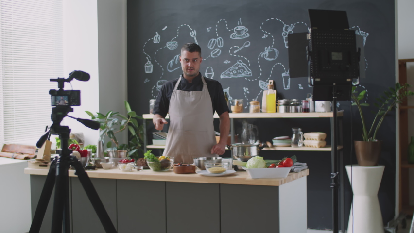 Caucasian man in apron presenting ingredients and describing recipe on camera while filming vlog for social media channel in kitchen Royalty-Free Stock Footage #1072579358