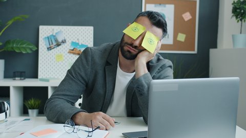 Inefficient office worker is sleeping at desk in workplace with sticky notes showing open eyes on face. Unmotivated employees and lazy people concept.