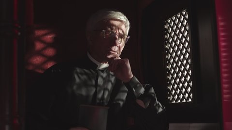 Medium shot of senior Caucasian priest wearing black robe, white collar and eyeglasses sitting in confessional near window, listening to confession and the reading Holy Bible