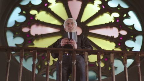 Low angle view of elderly Caucasian pastor wearing black clothes and white collar standing in Lutheran church against stained-glass window and preaching sermon