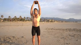 Shirtless man performing functional exercises with kettlebell on the beach at sunrise in slow motion
