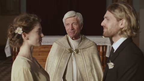 Medium shot of young Caucasian man and his beautiful bride standing at altar in Christian church during wedding ceremony while aged priest preaching sermon