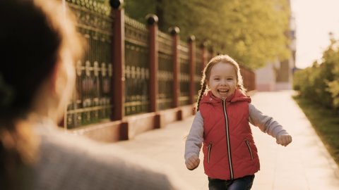 Child, daughter runs to mom and hugs her in park on street in spring. Happy family. Carefree childhood, joyful run of baby to mother. Small child cheerfully plays in summer on street with parent