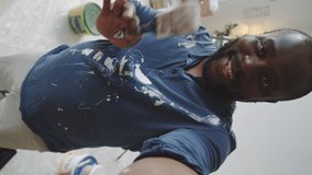 Vertical shot of cheerful Afro-American man in clothes with paint stains holding smartphone in outstretched arm, smiling and speaking at camera while showing home renovation on web call
