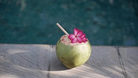 Footage of green young coconut close up with bamboo straw and tropical pink flower on the edge of swimming pool in Bali