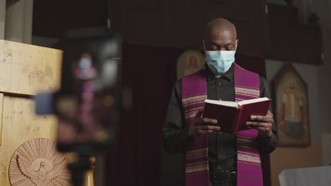 Racked focus shot of African-American pastor wearing medical mask standing in Lutheran church in front of smartphone with Holy Bible in hands and preaching sermon