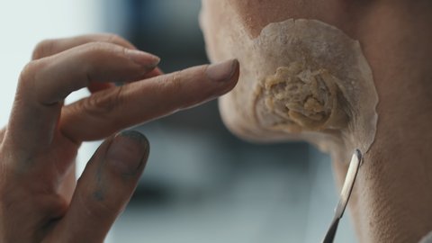 Close up of hands of unrecognizable female SFX makeup artist using spatula and applying prosthetic scar on man