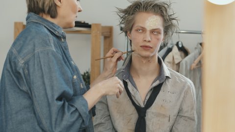 Slowmo tilt up shot of female SFX artist applying makeup on male actor with white contact lenses
