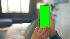 Woman at home lying on a bed and using smartphone with green mock-up screen in vertical mode. Girl browsing Internet, watching content, videos. POV.