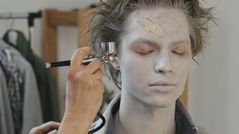 Close up shot of unrecognizable female SFX artist using airbrush machine and putting white makeup on face of male actor with prosthetic scar on forehead