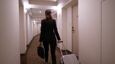 Business woman arrive to destination, check-in and walk to room and by long bending corridor. She carry small luggage, trolley case and laptop bag. Static camera, wide angle shot of passage