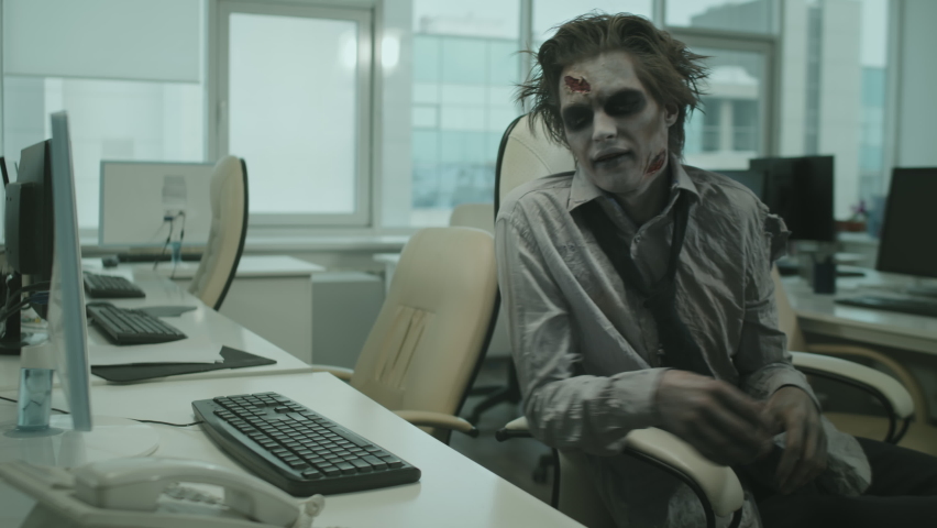 Medium shot of zombie office worker sitting at desk and smashing keyboard with his hands while trying to type