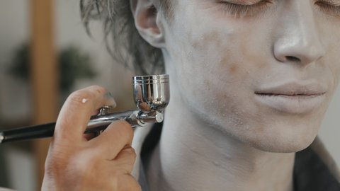 Handheld close up shot of unrecognizable female SFX artist using airbrush machine and applying scary makeup on young man