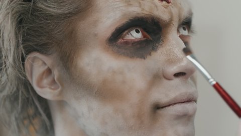 Close up shot of unrecognizable SFX artist applying black eyeshadow on eyes of young man with zombie makeup and white contact lenses