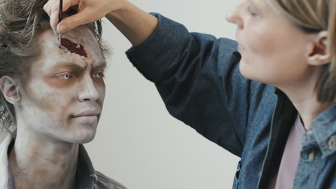 PAN shot of female SFX makeup artist creating fake wound on forehead of man with contact lenses and zombie makeup