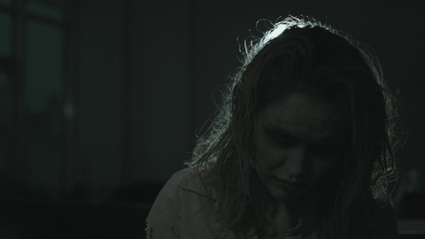 Medium shot of mindless female zombie with dirty hair, torn clothes and SFX makeup and contact lenses swaying and looking at camera in dark