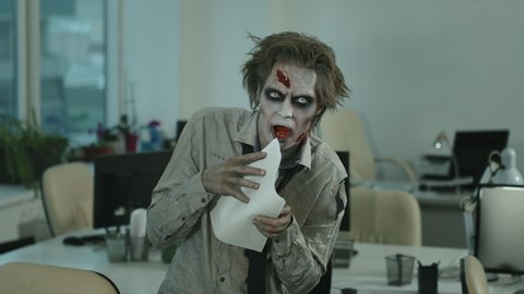 Portrait shot of zombie man with contact lenses, SFX makeup and fake wound on forehead looking at camera and eating document in office