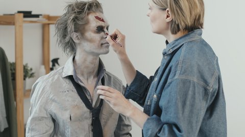 Medium shot of female SFX artist doing zombie makeup on man in torn shirt and necktie