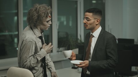 Medium shot of businessman in suit and necktie holding coffee cup and having conversation with zombie employee with SFX makeup and torn clothes
