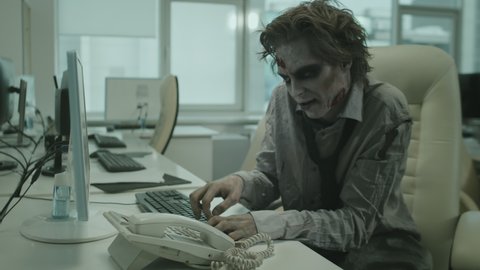 Handheld shot of zombie businessman with scary SFX makeup sitting at desk and screaming into phone while trying to make call