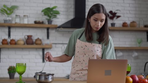Middle shot of smiling young slim Asian woman mixing sauce in cooking pan looking at laptop screen. Portrait of beautiful positive chef in apron preparing tasty homemade food indoors in kitchen