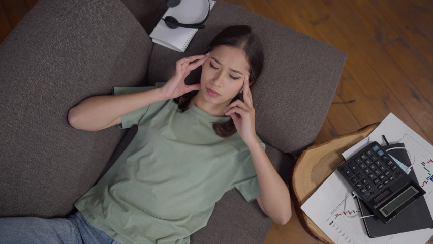 Top view portrait of overburdened exhausted young Asian woman lying on couch rubbing temples. Sad stressed tired freelancer having migraine symptoms in home office. Overworking and burnout Royalty-Free Stock Footage #1072600421