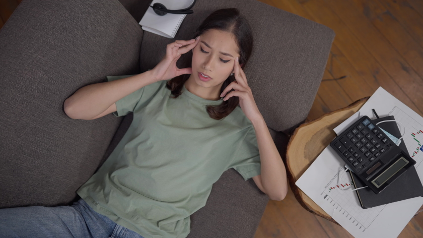 Top view portrait of overburdened exhausted young Asian woman lying on couch rubbing temples. Sad stressed tired freelancer having migraine symptoms in home office. Overworking and burnout | Shutterstock HD Video #1072600421