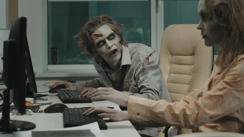 Medium shot with slowmo of mindless male and female zombies with SFX makeup and torn dirty clothes sitting at desks and smashing their hands on keyboard and mouse while trying to work on computers