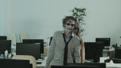 Slowmo shot of zombie businessman and businesswoman with SFX makeup and dirty clothes walking mindlessly in office