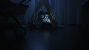 Two sisters play tablet at home. The girls are sitting in a wigwam and watching and touching the tablet screen. Video of happy children in the home room at night.