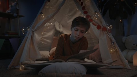 Teen boy in makeshift hut reading book at home in evening. Positive smart teenager lying on floor while spending free time. Concept of leisure and careless childhood