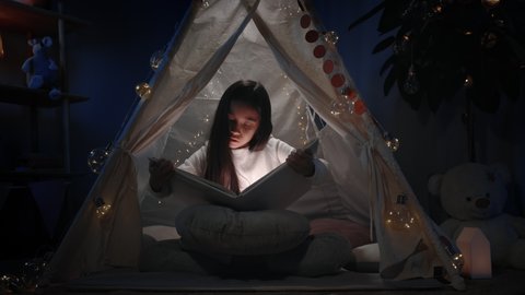 Beautiful girl with long black hair in decorative makeshift hut looking to book in evening. Pretty female teenager sitting on floor while reading . Concept of leisure and free time
