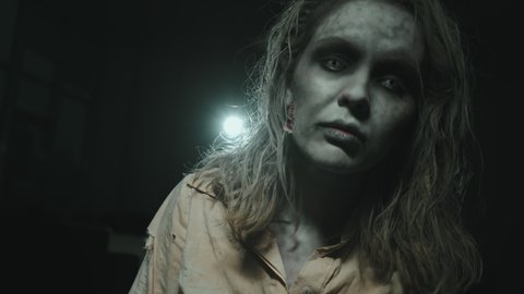 Close up shot of zombie woman with SFX makeup and contact lenses walking towards camera in darkness with spotlight shining in background