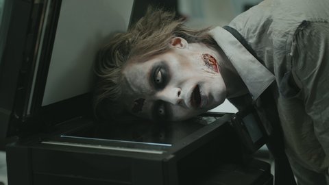 Medium shot of zombie man with SFX makeup and fake wounds grunting and photocopying his face on copier