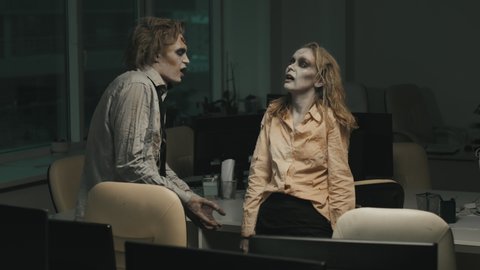 Medium shot with slowmo of grunting zombie man and woman with SFX makeup and torn dirty clothes having conversation in dark office
