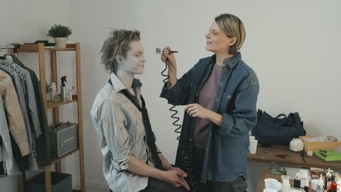 PAN shot of cheerful female SFX artist airbrushing white makeup on face of male horror movie actor