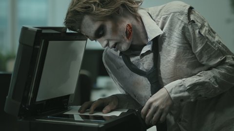 Medium shot of grunting zombie businessman in torn and dirty shirt and necktie and SFX makeup using copier in office
