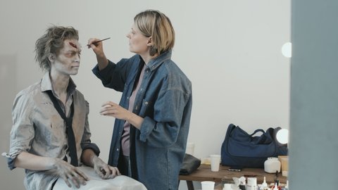 PAN of professional female makeup artist using brush and doing SFX zombie makeup on young man in torn shirt and necktie