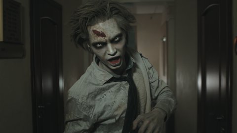 Tracking shot of creepy zombie man with SFX makeup and contact lenses running along dark hallway and following camera