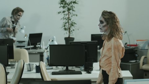 PAN slowmo of zombie businesswoman and businessman wearing SFX makeup, contact lenses and dirty clothes walking between desks in empty office