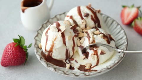 Vanilla ice cream scoops with chocolate topping and nuts, eating with spoon. Closeup view. Summer frozen dessert, sugar bomb