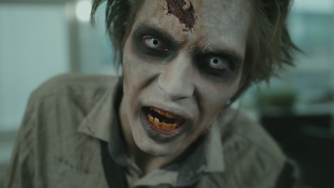 Handheld close up of zombie man with white contact lenses and SFX makeup with fake wounds looking at camera and grunting