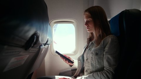 Young Woman Reads E-book while Flying in Airplane and Looking at Window. Touristic Leisure with E-reader or Tablet during Aircraft Flight in Economy Class. 4K Handheld Medium Shot