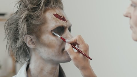 Close up shot of professional SFX makeup artist applying black eyeshadow on eyelids of man with zombie makeup and fake wound on his forehead