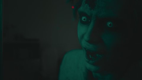 Close up shot of creepy zombie man with white contact lenses and SFX makeup twitching and grunting in darkness with red and blue lights flashing