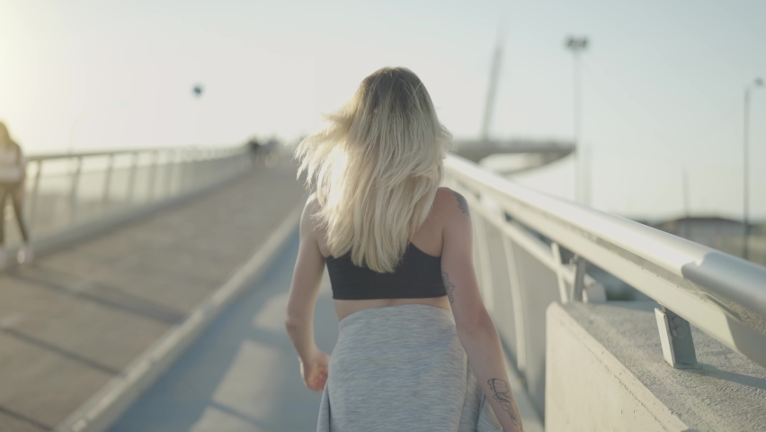Portrait back shot of a pretty young woman walking on a bridge in the city at sunset. Girl works out for wellbeing in summer.