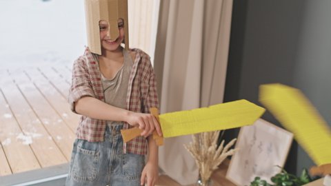 Slowmo of laughing 10-year-old girl in cardboard helmet fighting with toy swords with elder brother in modern countryside house