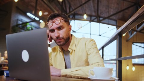Exhausted millennial male hold head with hand sitting at office table falling asleep, tired man fall asleep working at laptop at workplace, sleepy guy feel fatigue taking nap near computer