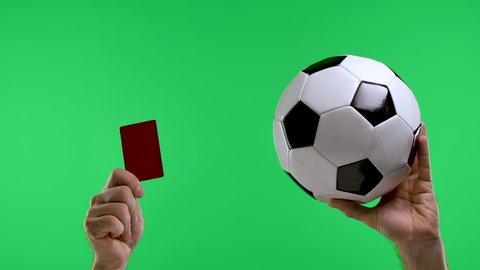 A male hand is holding a soccer ball and the second hand is showing a red card, isolated on a studio green screen of chromaticity key. Removing a player from the field. Slow motion. Close up.
